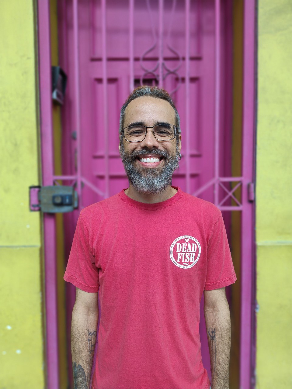 Dukão, palestrante do evento, homem sorrindo com cabelos e barba grizalhos, óculos e uma camisa rosa escrito Dead Fish